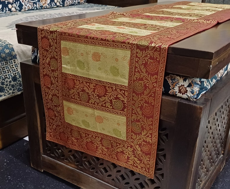 May include: A red and gold table runner with a floral pattern. The runner is made of a brocade fabric and has a fringed edge. The runner is on a wooden table with a carved design.