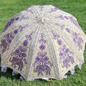May include: A white and purple umbrella with an embroidered design of elephants and flowers. The umbrella is closed and lying on a green grassy surface.