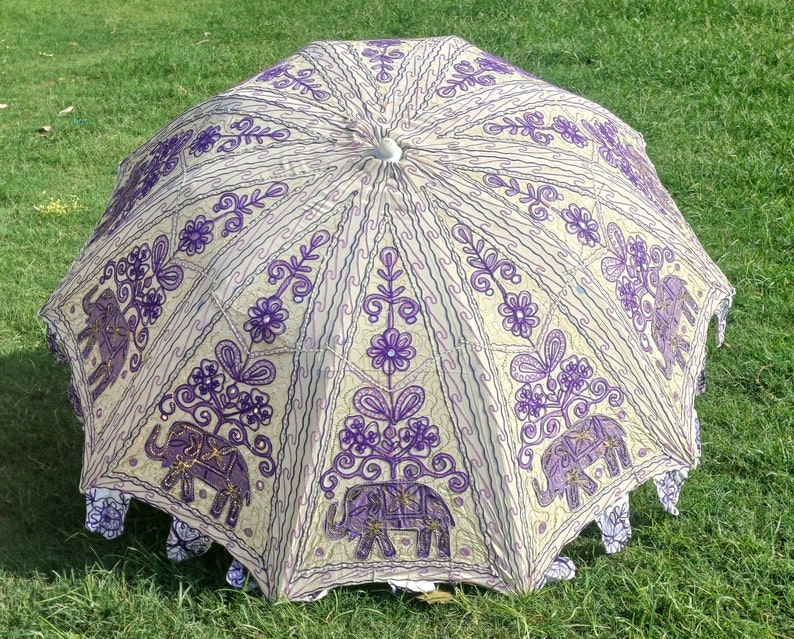 May include: A white and purple umbrella with an embroidered design of elephants and flowers. The umbrella is closed and lying on a green grassy surface.