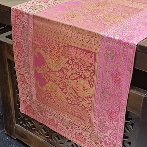 Puede incluir: Caminero de mesa rosa con un diseño de pavo real bordado en oro. El camino de mesa tiene un borde floral y está hecho de una tela suave y lujosa.