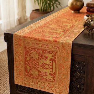 May include: A rectangular table runner in a vibrant orange hue featuring intricate gold-toned woven patterns. The design displays repeating motifs of elephants and floral elements, framed by a detailed border with geometric and botanical accents.