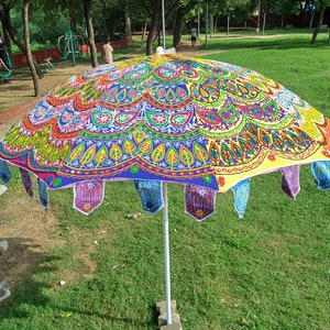 May include: A large, round garden umbrella featuring a vibrant, multi-colored fabric canopy with intricate embroidered patterns, including floral motifs and elephant shapes along the scalloped edge, supported by a central white metal pole.