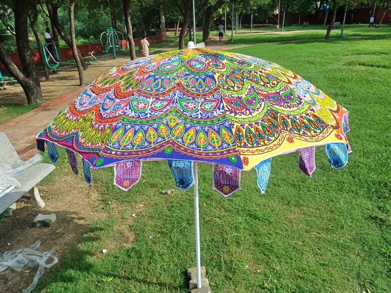 May include: A large, round garden umbrella featuring a vibrant, multi-colored fabric canopy with intricate embroidered patterns, including floral motifs and elephant shapes along the scalloped edge, supported by a central white metal pole.