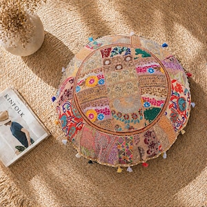 Könnte beinhalten: Runder Bodenkissen mit Patchwork-Design in Beige-, Rosa- und Blautönen. Das Kissen hat einen dekorativen Rand und ist mit kleinen Quasten verziert. Eine Zeitschrift und eine Vase befinden sich im Hintergrund.