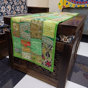 K&ouml;nnte beinhalten: Ein gr&uuml;ner Patchwork-Tischl&auml;ufer mit Stickereien, der eine Mischung aus floralen und geometrischen Mustern aufweist. Der L&auml;ufer liegt auf einem dunklen Holztisch mit geschnitzten Details.