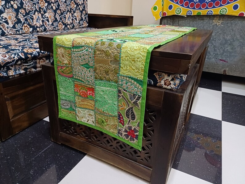 K&ouml;nnte beinhalten: Ein gr&uuml;ner Patchwork-Tischl&auml;ufer mit Stickereien, der eine Mischung aus floralen und geometrischen Mustern aufweist. Der L&auml;ufer liegt auf einem dunklen Holztisch mit geschnitzten Details.
