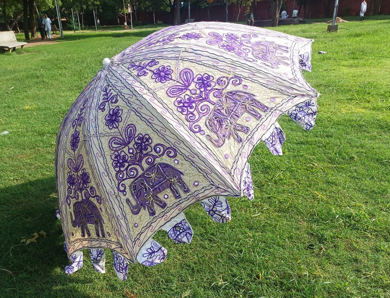May include: A white and purple umbrella with an embroidered elephant design. The umbrella is open and has a fringe border.
