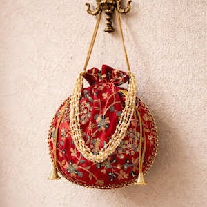 May include: A round, red velvet bag with intricate floral embroidery in gold, green, and blue. The bag features a drawstring closure and is adorned with a pearl necklace. Gold tassels hang from the bottom, and the bag is suspended from a decorative gold hook.