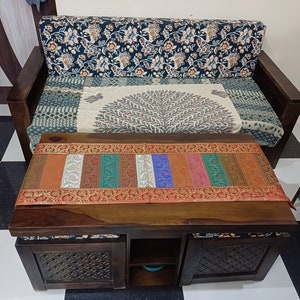 以下が含まれることがあります： カラフルな模様のランナーと模様入りのクッションが付いた木製ソファを備えた木製のコーヒーテーブル。 テーブルには収納コンパートメントがあり、白黒の市松模様の床に置かれています。