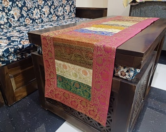 May include: A pink, gold and green table runner with a floral pattern. The runner is on a dark wood table with carved details.