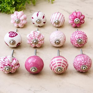 Peut inclure: Un ensemble de douze boutons de placard décoratifs dans des tons rose et blanc. Chaque bouton présente une forme ronde ou florale avec des motifs variés tels que des rayures, des pois, des cœurs et des dessins botaniques, avec une tige métallique.