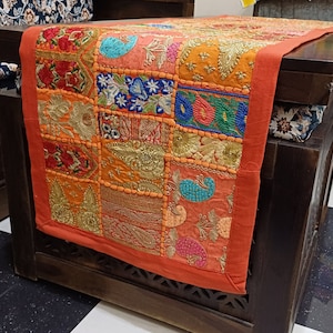 May include: Orange table runner with a patchwork design featuring colorful embroidered floral patterns and gold accents. The runner is made of fabric and has a fringed edge.