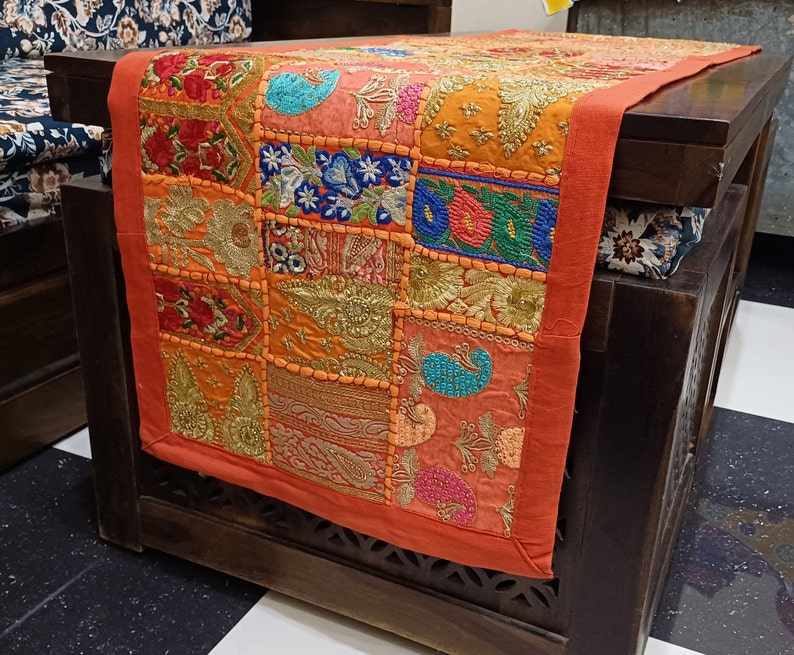 Puede incluir: Un camino de mesa naranja con un dise&ntilde;o de patchwork que presenta coloridos patrones florales bordados y detalles dorados. El camino de mesa est&aacute; hecho de tela y tiene un borde con flecos.