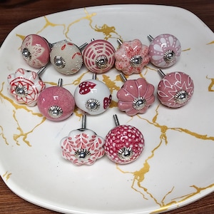 K&ouml;nnte beinhalten: Eine Sammlung von elf runden Keramik-Schrankkn&ouml;pfen mit verschiedenen rosa, wei&szlig;en und roten Mustern, darunter Blumen-, Punkte- und Herzdesigns. Jeder Knopf hat eine mittige silberfarbene Metallschraube und eine polierte silberfarbene Kappe.