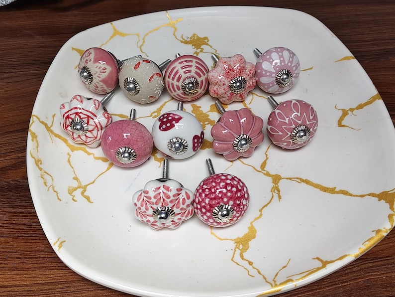 K&ouml;nnte beinhalten: Eine Sammlung von elf runden Keramik-Schrankkn&ouml;pfen mit verschiedenen rosa, wei&szlig;en und roten Mustern, darunter Blumen-, Punkte- und Herzdesigns. Jeder Knopf hat eine mittige silberfarbene Metallschraube und eine polierte silberfarbene Kappe.