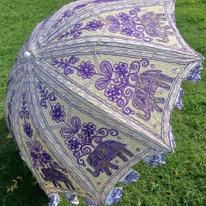May include: A white and purple umbrella with an embroidered design of elephants and flowers. The umbrella is made of cotton and has a fringe border.