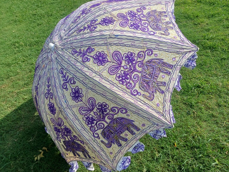 May include: A white and purple umbrella with an embroidered design of elephants and flowers. The umbrella is made of cotton and has a fringe border.
