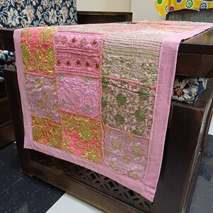 K&ouml;nnte beinhalten: Ein rosa Patchwork-Tischl&auml;ufer mit goldener Stickerei und einem Fransenrand. Der L&auml;ufer besteht aus verschiedenen Stoffen und Texturen und hat einen einzigartigen und bohemischen Stil.