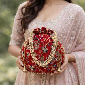 May include: A round, red velvet clutch with floral embroidery and a beaded strap. The bag features gold tassels and a drawstring closure. The design is ornate and elegant, suitable for special occasions.