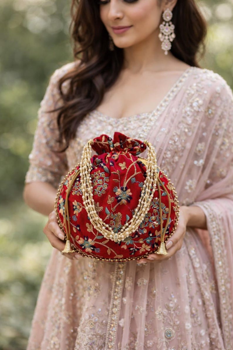 May include: A round, red velvet clutch with floral embroidery and a beaded strap. The bag features gold tassels and a drawstring closure. The design is ornate and elegant, suitable for special occasions.
