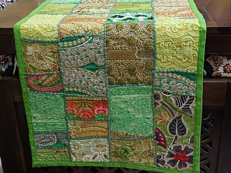 K&ouml;nnte beinhalten: Ein gr&uuml;ner Patchwork-Tischl&auml;ufer mit Stickereien. Der L&auml;ufer besteht aus verschiedenen Stoffen und zeigt eine Vielzahl von Farben, darunter Gr&uuml;n, Gold und Rosa.