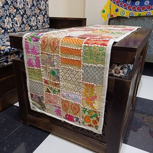 K&ouml;nnte beinhalten: Ein wei&szlig;er Tischl&auml;ufer mit Patchwork-Design, das aus bunten, bestickten Quadraten besteht. Der L&auml;ufer ist aus Baumwolle und hat einen Fransenrand. Er wird auf einem Holztisch mit dunkelbrauner Oberfl&auml;che gezeigt.