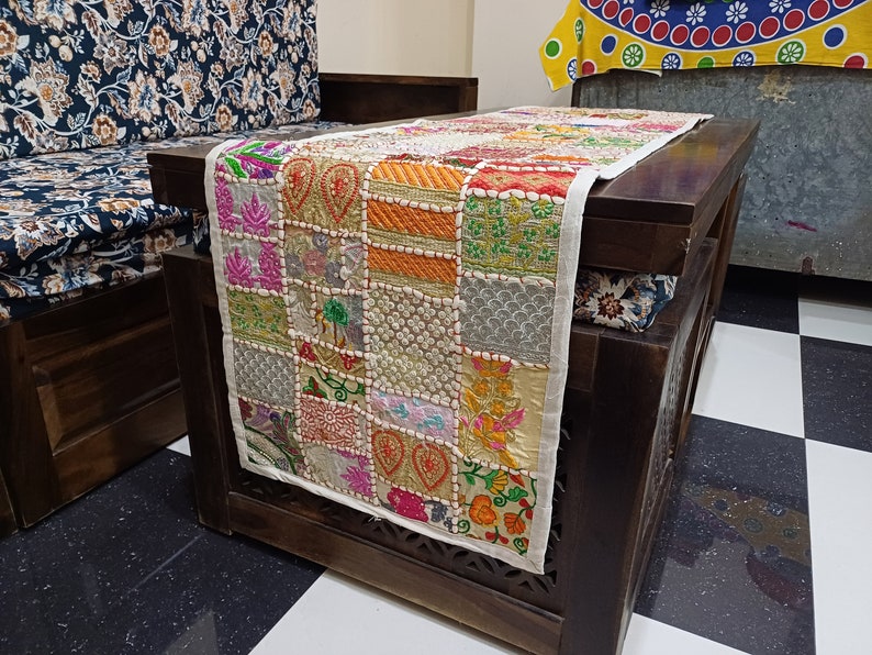 K&ouml;nnte beinhalten: Ein wei&szlig;er Tischl&auml;ufer mit Patchwork-Design, das aus bunten, bestickten Quadraten besteht. Der L&auml;ufer ist aus Baumwolle und hat einen Fransenrand. Er wird auf einem Holztisch mit dunkelbrauner Oberfl&auml;che gezeigt.