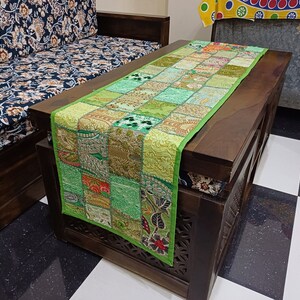 K&ouml;nnte beinhalten: Gr&uuml;ner Patchwork-Tischl&auml;ufer mit goldenen und silbernen Stickereien. Der L&auml;ufer liegt auf einem dunklen Holztisch mit geschnitzten Details. Der Tisch steht vor einem Sofa mit einem floralen Stoffmuster.