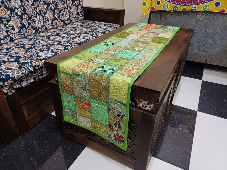 K&ouml;nnte beinhalten: Gr&uuml;ner Patchwork-Tischl&auml;ufer mit goldenen und silbernen Stickereien. Der L&auml;ufer liegt auf einem dunklen Holztisch mit geschnitzten Details. Der Tisch steht vor einem Sofa mit einem floralen Stoffmuster.