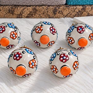 May include: Five round ceramic cabinet knobs with a cream-colored base. Each knob features a hand-painted floral pattern with red, blue, and orange petals, black stems, and a prominent, solid orange circular center piece on the front.
