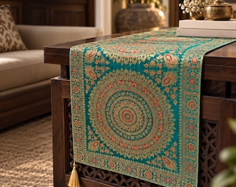 Puede incluir: Un camino de mesa rectangular de color verde azulado con intrincados patrones de mandalas circulares y motivos florales en oro y rojo. El camino está rematado con dos borlas de color oro claro que cuelgan de las esquinas inferiores.