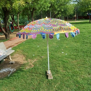May include: A round outdoor umbrella featuring a vibrant, multi-colored mandala pattern in shades of yellow, pink, green, and blue. The canopy edge is decorated with small, dangling fabric pennants in various colors, supported by a central pole.