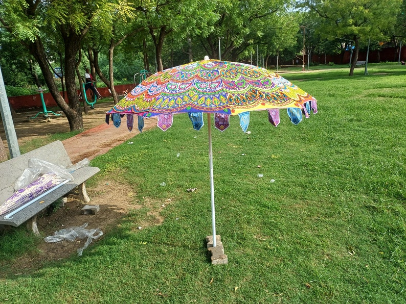 May include: A round outdoor umbrella featuring a vibrant, multi-colored mandala pattern in shades of yellow, pink, green, and blue. The canopy edge is decorated with small, dangling fabric pennants in various colors, supported by a central pole.