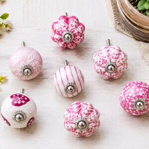 K&ouml;nnte beinhalten: Eine Sammlung von sieben Keramik-M&ouml;belkn&ouml;pfen in verschiedenen Rosa- und Wei&szlig;t&ouml;nen. Die Kn&ouml;pfe weisen unterschiedliche Muster wie Streifen, florale Motive und Punkte auf, jeweils mit einem zentralen Beschlag aus silberfarbenem Metall.