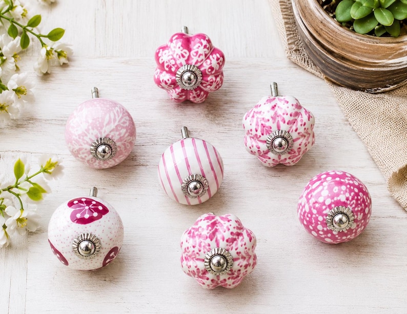 K&ouml;nnte beinhalten: Eine Sammlung von sieben Keramik-M&ouml;belkn&ouml;pfen in verschiedenen Rosa- und Wei&szlig;t&ouml;nen. Die Kn&ouml;pfe weisen unterschiedliche Muster wie Streifen, florale Motive und Punkte auf, jeweils mit einem zentralen Beschlag aus silberfarbenem Metall.