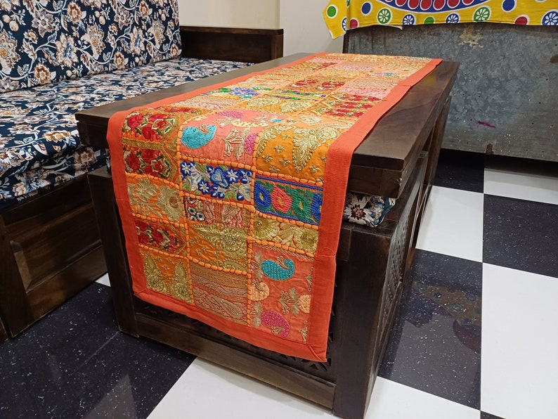 Puede incluir: Un camino de mesa de patchwork colorido con acentos naranja, rojo y dorado. El camino de mesa est&aacute; hecho de cuadrados de tela cosidos juntos y presenta intrincados detalles de bordado.