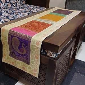 May include: A table runner with a cream background and a colorful patterned design. The pattern features a peacock, floral motifs, and geometric shapes in shades of purple, pink, red, orange, yellow, and green. The runner is placed on a dark wooden table.