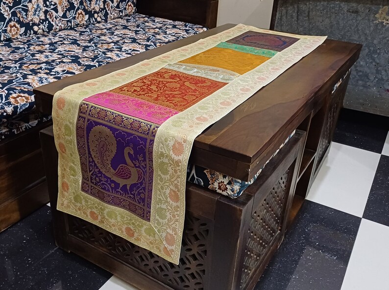 May include: A table runner with a cream background and a colorful patterned design. The pattern features a peacock, floral motifs, and geometric shapes in shades of purple, pink, red, orange, yellow, and green. The runner is placed on a dark wooden table.