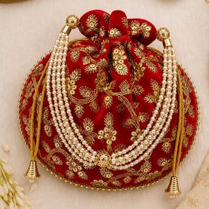 Può includere: Una pochette rotonda in velluto rosso con chiusura a coulisse, caratterizzata da ricami metallici dorati e decorazioni con paillettes. La borsa è arricchita da tre fili di perle bianche artificiali su ogni lato, con terminali conici dorati.