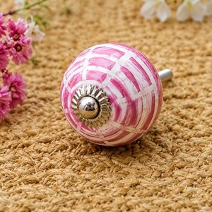 K&ouml;nnte beinhalten: Ein runder Keramik-M&ouml;belknopf mit einem handgemalten rosa-wei&szlig;en Gittermuster. Die Mitte des Knopfes ist mit einer silberfarbenen Metallkappe mit gewelltem Rand verziert, und ein silberfarbener Metallbolzen ragt aus der R&uuml;ckseite des Knopfes heraus.