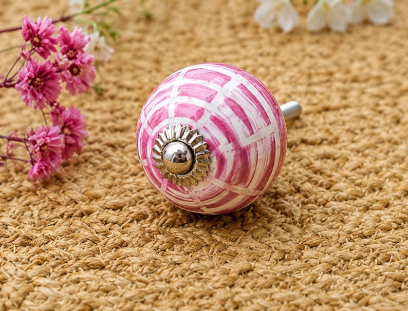 K&ouml;nnte beinhalten: Ein runder Keramik-M&ouml;belknopf mit einem handgemalten rosa-wei&szlig;en Gittermuster. Die Mitte des Knopfes ist mit einer silberfarbenen Metallkappe mit gewelltem Rand verziert, und ein silberfarbener Metallbolzen ragt aus der R&uuml;ckseite des Knopfes heraus.