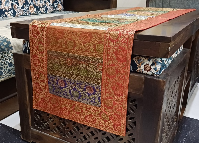 以下が含まれることがあります： 赤、金、緑、紫の色合いの花柄が施された長方形のテーブルランナー。 ランナーは濃い色の木製テーブルの上に掛けられています。 ランナーの端は、詳細な花柄のデザインで縁取られています。