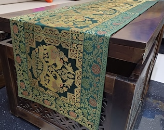 Puede incluir: Camino de mesa verde y dorado con diseño de elefantes. El corredor presenta un patrón detallado con elefantes, motivos florales y bordes decorativos. La tela parece tejida, agregando textura. Se coloca sobre una mesa de madera oscura.