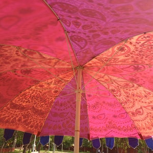 May include: A large outdoor umbrella featuring a vibrant canopy with alternating panels of pink, orange, and red, decorated with intricate paisley and mandala-inspired patterns. The umbrella has a central pole and dark blue fabric fringe along the edge.