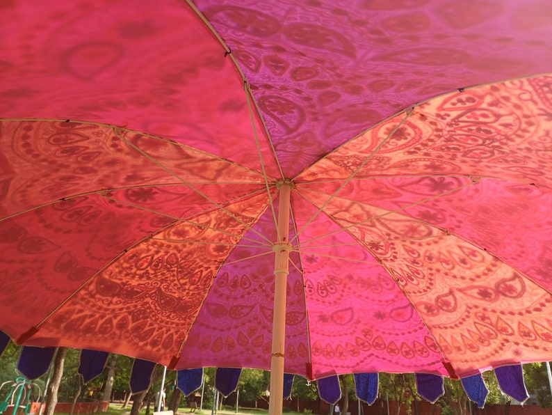 May include: A large outdoor umbrella featuring a vibrant canopy with alternating panels of pink, orange, and red, decorated with intricate paisley and mandala-inspired patterns. The umbrella has a central pole and dark blue fabric fringe along the edge.