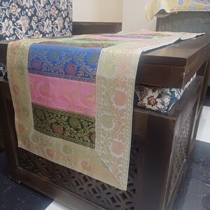 May include: A wooden table with a decorative table runner. The table runner is made of fabric with a multi-colored floral pattern. The table runner has a gold border.