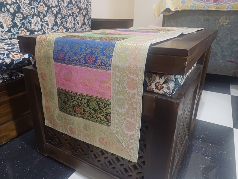 May include: A wooden table with a decorative table runner. The table runner is made of fabric with a multi-colored floral pattern. The table runner has a gold border.