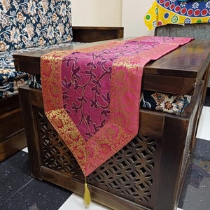 May include: A rectangular table runner in vibrant magenta featuring a dark purple vine pattern and wide, ornate gold metallic borders with floral motifs. The runner ends in a pointed tip adorned with a single, thin, light gold decorative tassel.