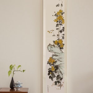 Puede incluir: Una pintura tradicional china en rollo que representa una rama de crisantemos amarillos creciendo desde una roca. El rollo está montado sobre un fondo blanco y está colgado en una pared blanca. Una pequeña mesa de madera con un jarrón y una taza de té se encuentra en primer plano.
