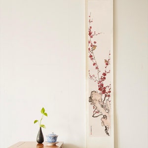 Puede incluir: Una pintura tradicional china en rollo que representa una rama delicada de flores de ciruelo con pequeños pájaros posados en las ramas. El rollo está montado sobre un fondo blanco y cuelga de una pared blanca. Una pequeña mesa de madera con un jarrón y una taza de té se encuentra en primer plano.